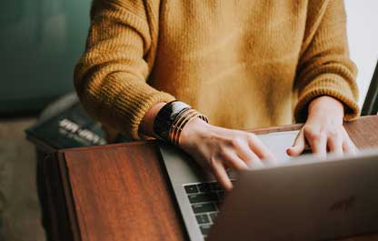 The hands and torso of a person sitting at a wooden desk wearing a knitted jumper, working on a laptop