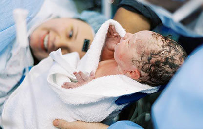 A photo of a newborn baby being held while her mum looks at her whilst in the delivery room.